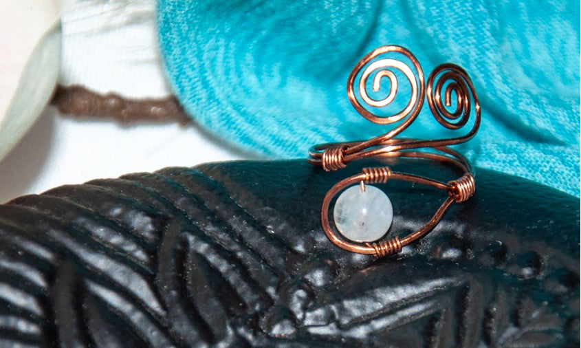 A handcrafted copper wire ring with a double spiral flourish and a wire-wrapped round moonstone bead at the base, resting on a dark teal ceramic surface with driftwood and a white magnolia flower in the background.