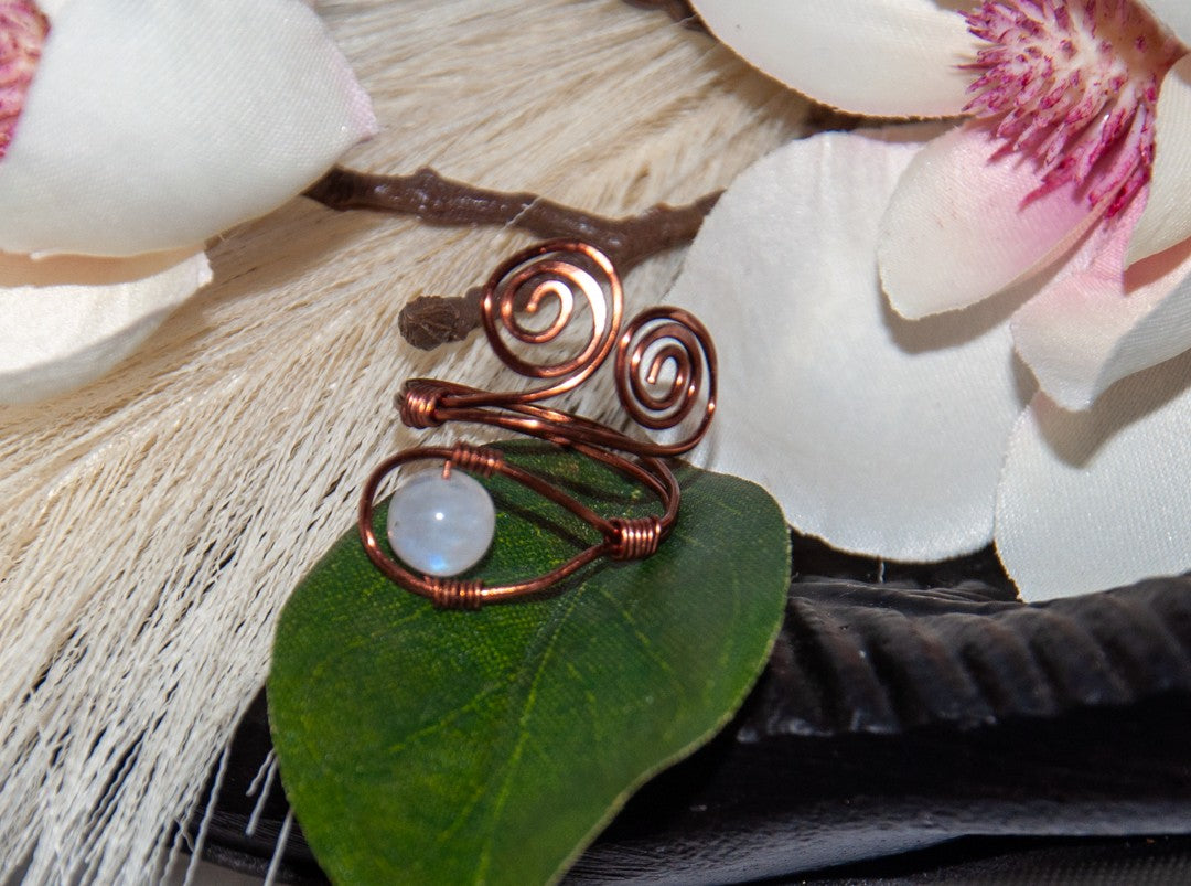 A handcrafted copper wire ring with a double spiral flourish and a wire-wrapped round moonstone bead at the base, resting on a green leaf with white magnolia flowers and driftwood in the background.