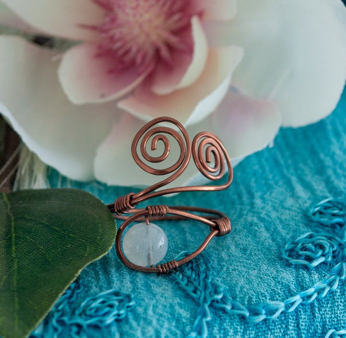 A handcrafted copper wire ring with a double spiral flourish and a wire-wrapped round moonstone bead at the base, displayed on a teal embossed surface with a soft-focus white and pink magnolia flower and green leaf in the background.